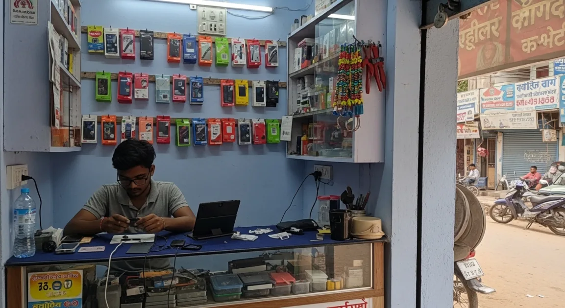 एक छोटे शहर की मोबाइल रिपेयरिंग दुकान जहाँ तकनीशियन स्मार्टफोन ठीक कर रहा है और colorful मोबाइल एक्सेसरीज़ लगी हैं।